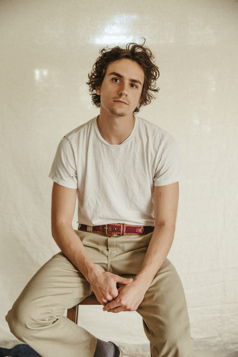 Male with dark curly hair perched on a stool wearing neutral coloured t-shirt and trousers by YAXA
